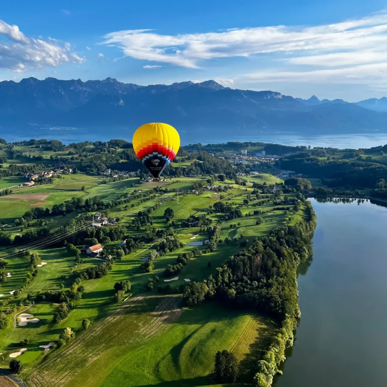 Vue d’une montgolfière survolant la campagne et un lac, avec le lac Léman et la chaîne des Alpes au loin